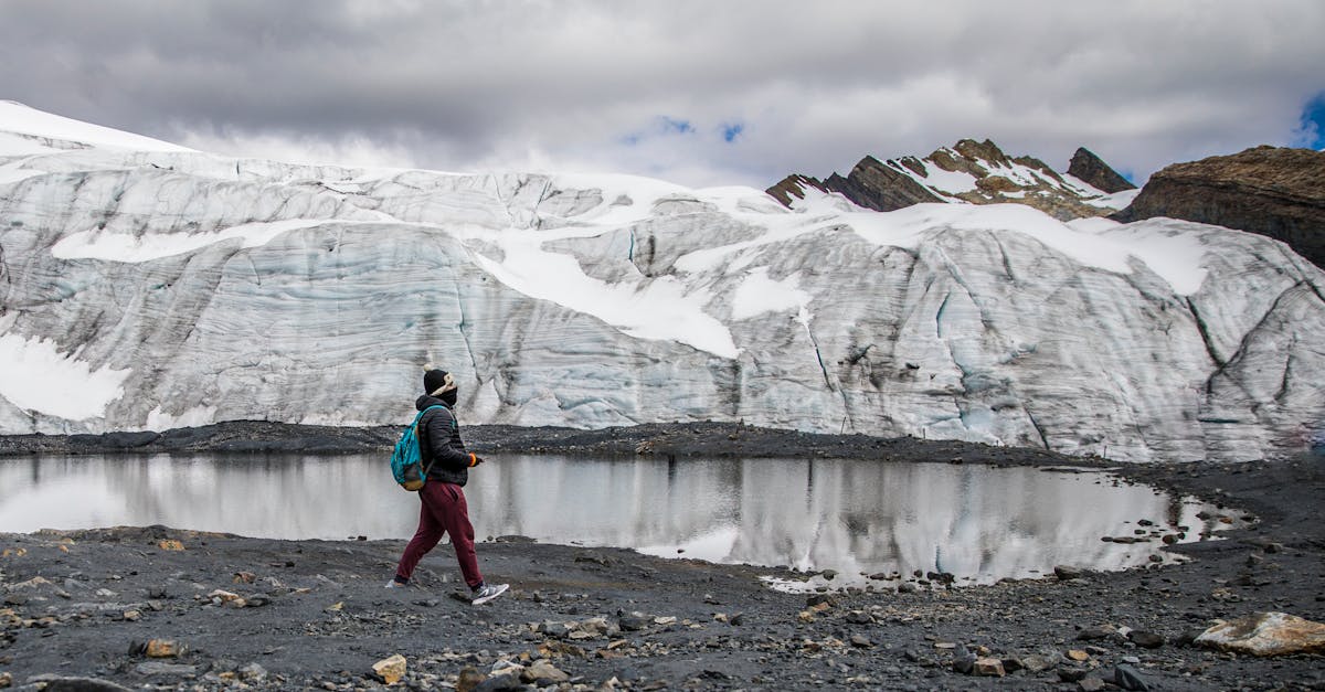 A lone hiker walks beside a glacier and a reflective pond under cloudy skies