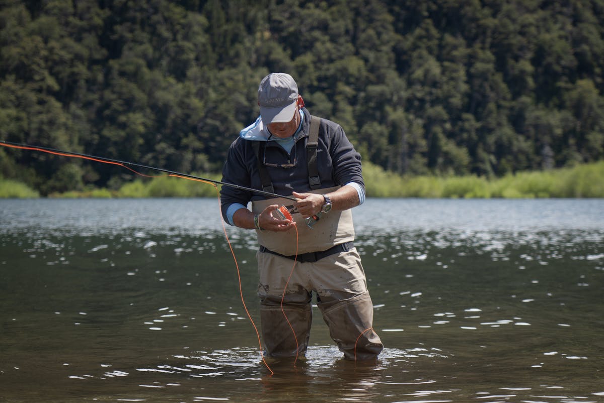 Angler wading in a calm Patagonian lake with forest-covered mountains in background