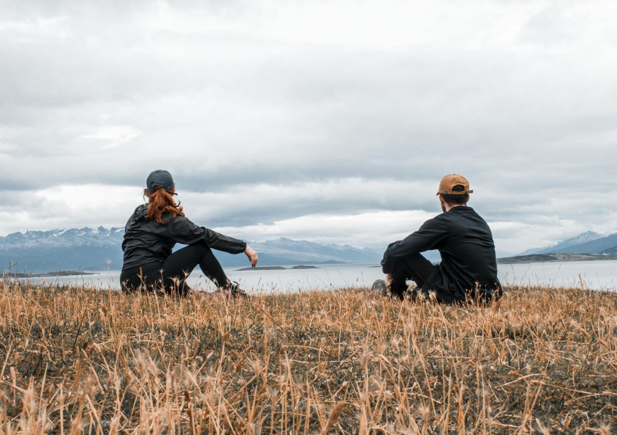 Couple sitting together on grass overlooking a lake surrounded by mountains