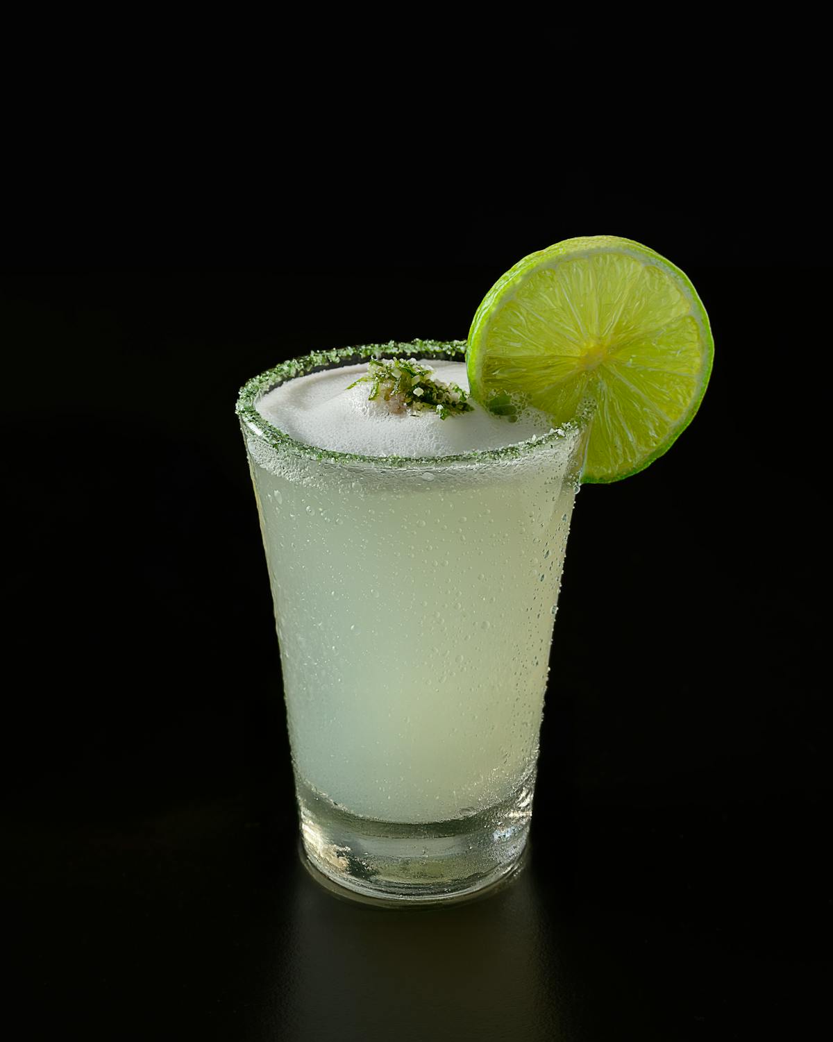 A pisco sour cocktail with a lime slice and sugar rim against a dark background
