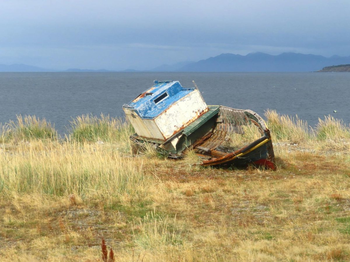 Weathered boat on grassy shore with ocean and mountains in the background at Punta Arenas